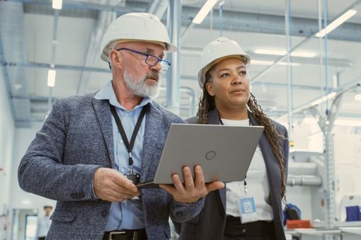 Two people talking while holding a computer and walking through a warehouse - Applied CAx Siemens machining manufacturing software